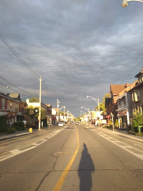 Image of a street in the sunlight against a cloudy, moody sky. The street is empty mostly, and stretched to the horizon. The shadow of the photographer stretches long in the bottom of the shot. 