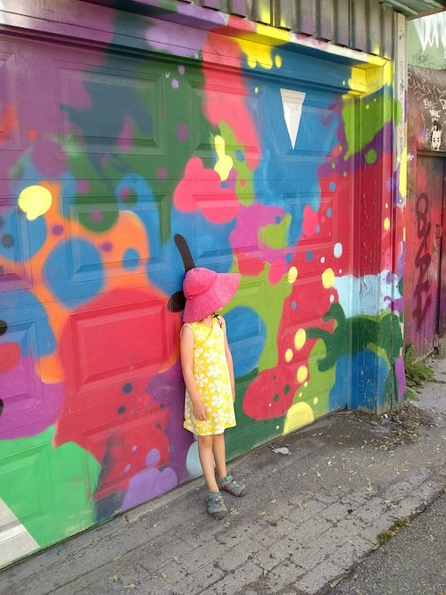 Image of a child in a yellow dress against a colourful background. She is wearing a big red hat and looking away from the camera so the viewer can't see her face. 