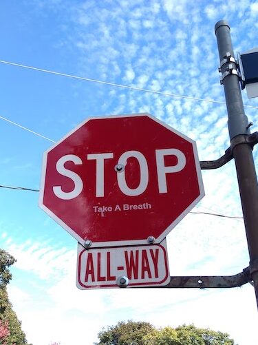 Photo of a STOP sign against a blue sky with some scattered clouds. Underneath the STOP, someone has pasted a stick that says "Take A Breath" 